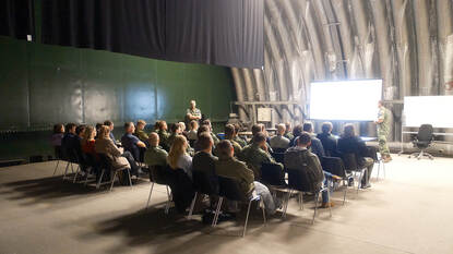 Twee militairen spreken een zaal vol mensen toe.