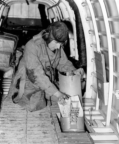 Zwart-witfoto van een bemanningslid dat op zijn knieën in een vliegtuig zit en een stapel pamfletten in een buis schuift.