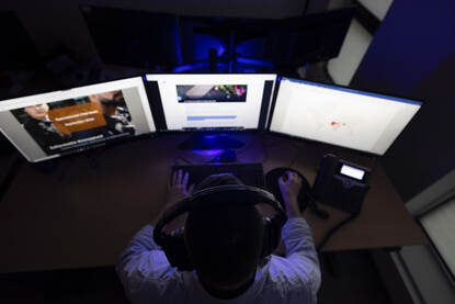 Persoon met hoofdtelefoon op zit aan een bureau met telefoon achter 3 beeldschermen.