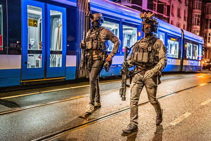 Twee medewerkers van de Dienst Speciale Interventies lopen zwaarbewapend naast elkaar. Daarachter een tram.