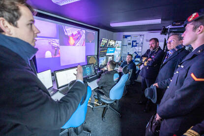 Genodigden kijken mee naar grote schermen met beelden van de zeebodem bij Plymouth.