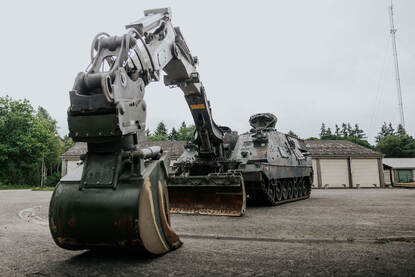 Een geparkeerde Kodiak. Gezien vanaf de voorzijde. De graafarm rust op de grond. Achter het geniedoorbraaksysteem zijn loodsen te zien.