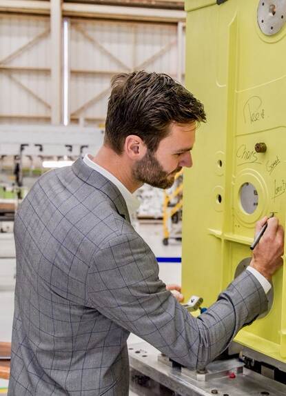 Een man in een net pak en een pen in zijn rechter hand schrijft iets op een groot geel metalen vlak.