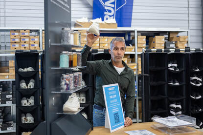 Links een man in vlekkenpak spreekt mannen in een oranje trainingsjack toe. Rechts een man die een witte schoen omhoog houdt.