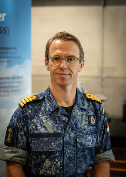 Een marineman in tenue met bril staat voor een groot bord.